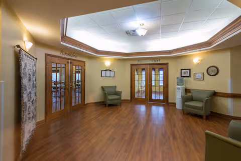 Interior view of a senior living facility common area with wooden flooring, beige walls, and a recessed ceiling light fixture. There are two sets of double wooden doors labeled 'Florentine' and 'Walnut Street'. The room contains three green armchairs, a water cooler, a clock, and framed pictures on the walls.