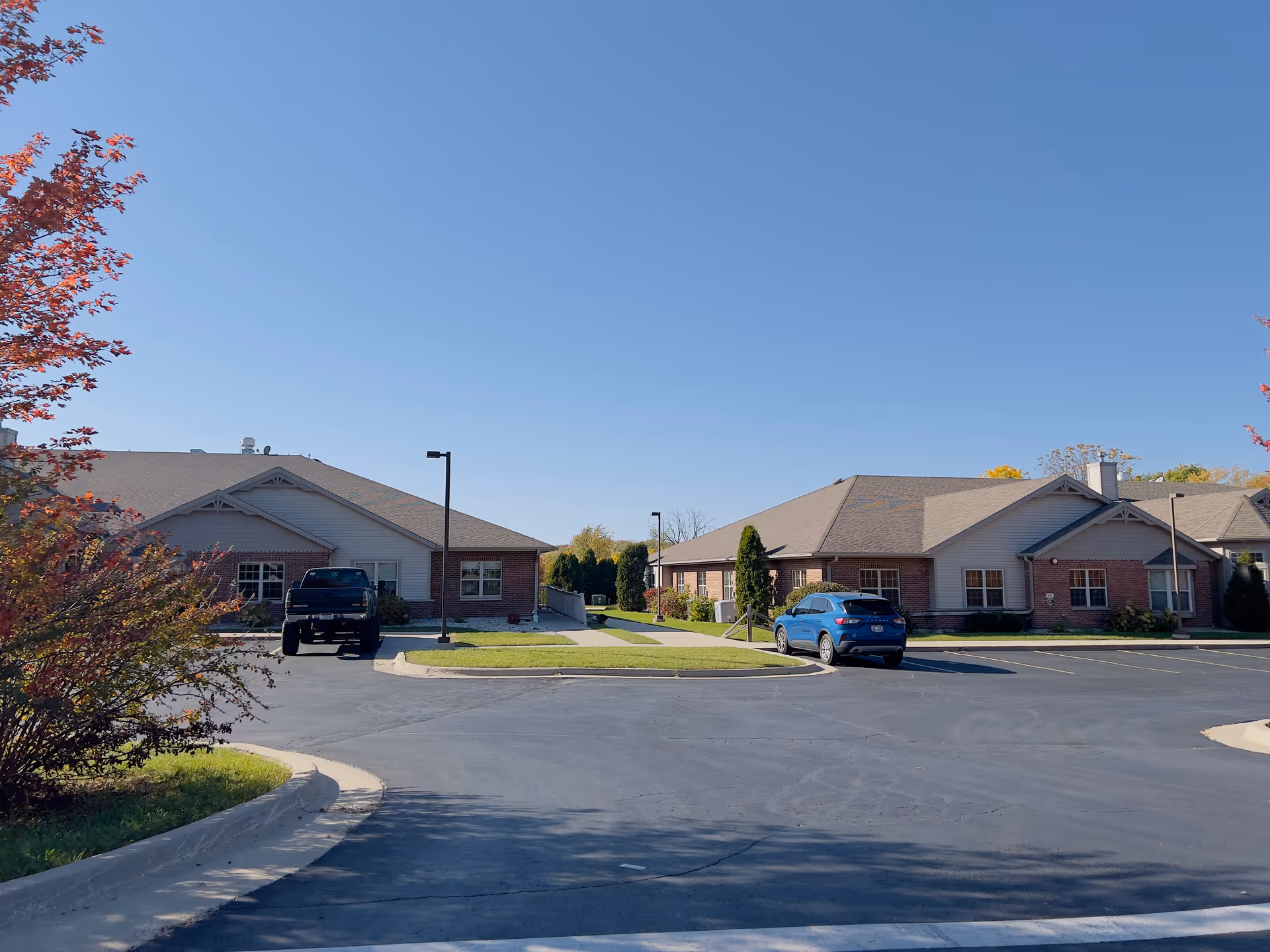 Low-rise brick senior living buildings with a parking lot, a few parked cars, and a clear blue sky.