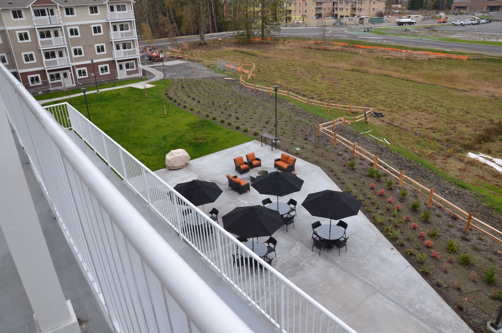 Aerial view of a courtyard patio with tables, black umbrellas and orange lounge chairs beside a multi-story residential building and landscaped lawn.