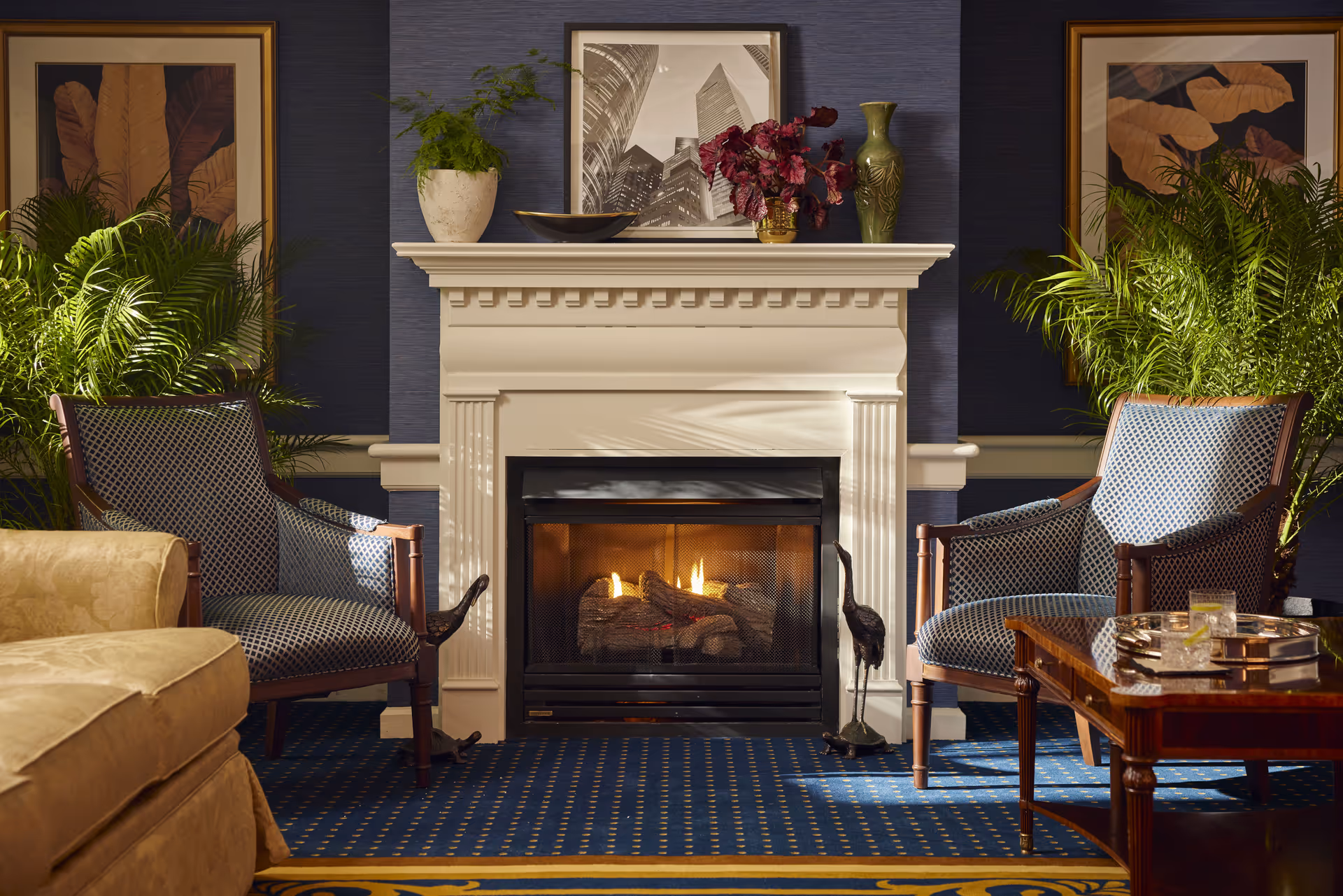 A cozy living room area featuring a lit fireplace with a white mantel. On the mantel are decorative items including a potted plant, a black and white framed photo of skyscrapers, a green vase, and a small bouquet of red flowers. Two patterned armchairs flank the fireplace, with green plants behind them. A wooden coffee table with a silver tray holding two glasses is in front of one chair. The room has a blue carpet with a yellow border and dark blue walls with framed botanical artwork.