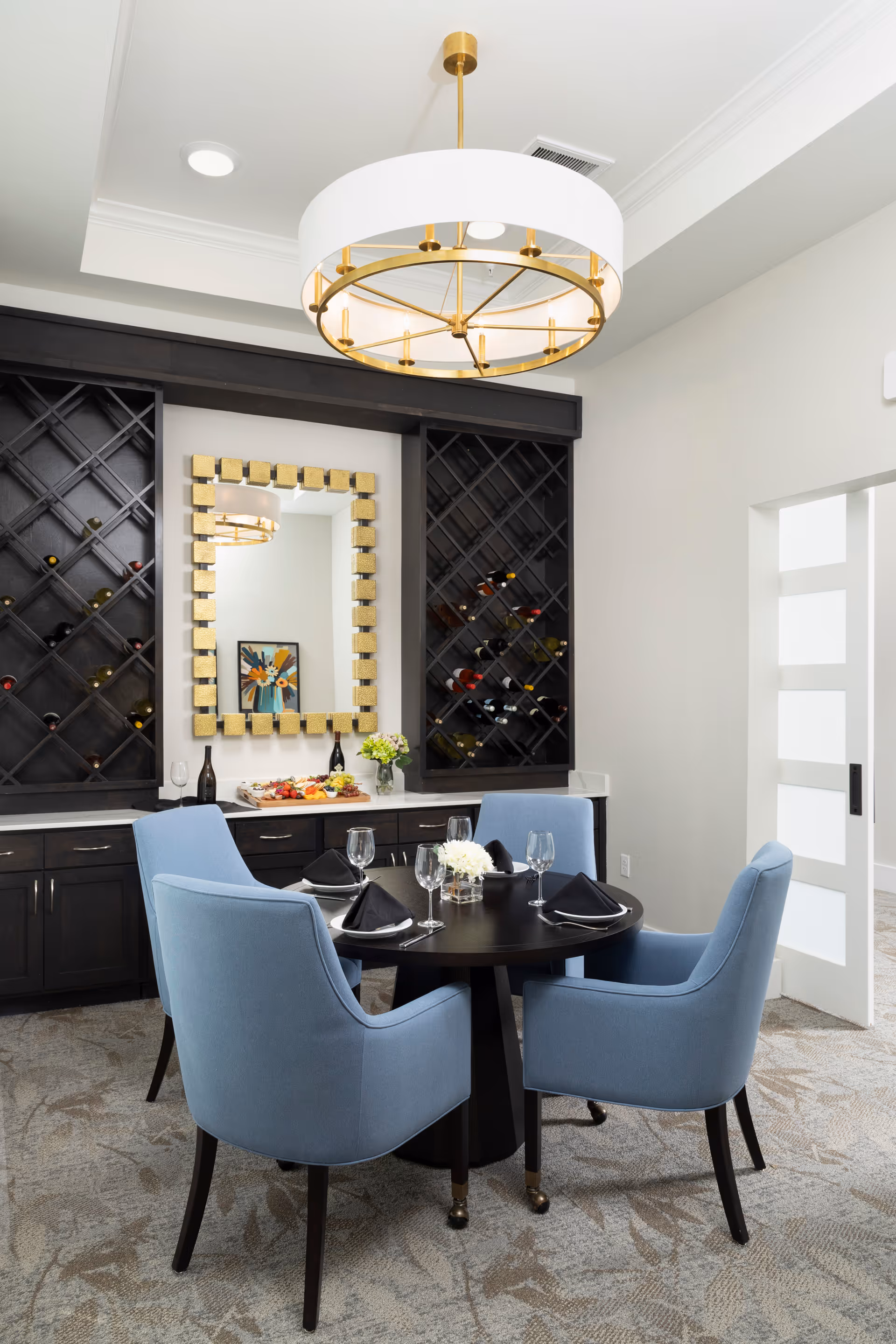 A dining area with a round black table set for four with black napkins and glassware. Four light blue upholstered chairs surround the table. Behind the table is a dark wood cabinet with built-in wine racks filled with bottles, a gold-framed mirror, and a tray with wine bottles and snacks. A modern chandelier with a white drum shade and gold accents hangs above the table. The room has light-colored walls and carpeted flooring with a subtle pattern.