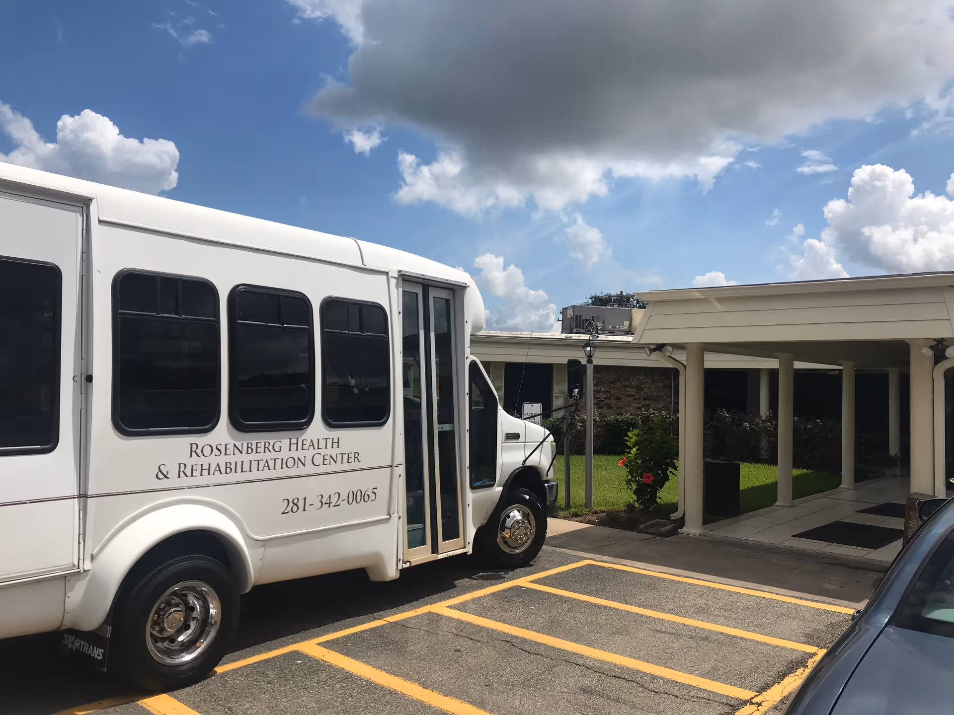A white shuttle bus parked outside the entrance of Rosenberg Health & Rehabilitation Center under a partly cloudy sky. The building entrance has a covered walkway supported by columns, and there is a small garden area with green grass and plants near the entrance.