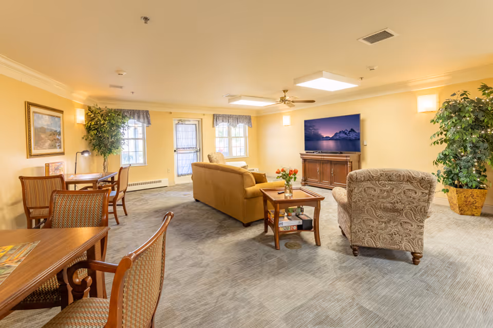 A cozy living room area in Brookridge Heights featuring a beige sofa, a patterned armchair, a wooden coffee table with a vase of flowers, a flat-screen TV on a wooden stand, two large potted plants, and a small dining area with tables and chairs near windows with curtains.