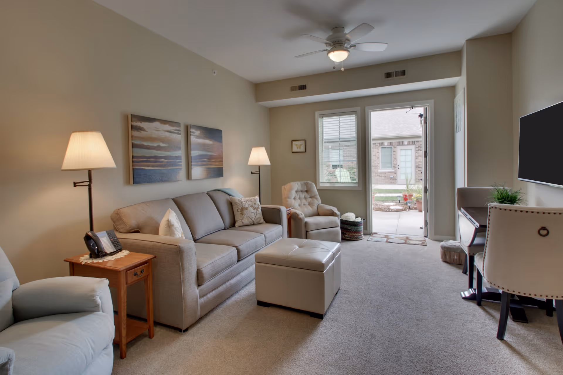 A cozy living room with beige walls and carpet, featuring a gray sofa with pillows, two armchairs, a small wooden side table with a phone and lamp, a ceiling fan with light, a wall-mounted TV, and a door leading to an outdoor patio area.