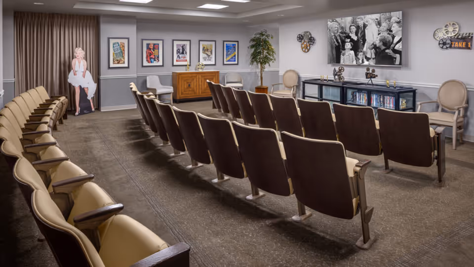 A small theater room with rows of beige and brown theater seats facing a large TV screen displaying a black and white movie scene. The room has movie posters on the wall, a wooden cabinet, a potted plant, and a life-size cutout of Marilyn Monroe near a curtain.