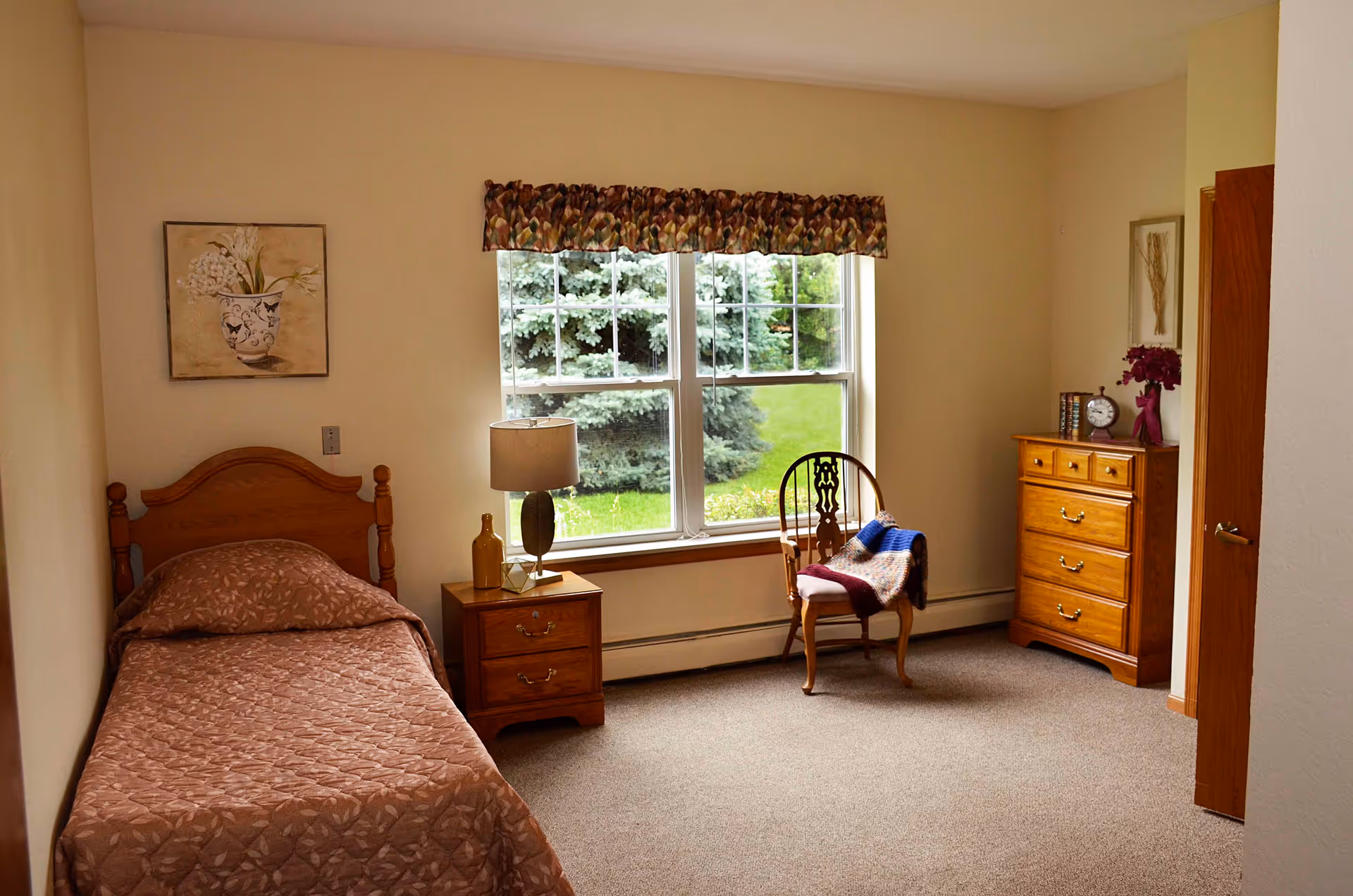 A simple bedroom with a single wooden bed covered with a brown patterned bedspread. Next to the bed is a wooden nightstand with a lamp and a decorative bottle. A wooden chair with a colorful knitted blanket is placed near a large window with a floral valance, showing green trees and grass outside. On the right side of the room is a wooden dresser with a clock, books, and a vase with flowers. The walls are light-colored, and the floor is carpeted.