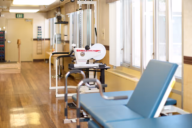 A rehabilitation or physical therapy room with exercise equipment including a stationary arm bike and parallel bars, wooden flooring, and large windows letting in natural light.