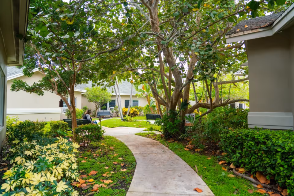 A curved concrete walkway winds through a shaded, landscaped courtyard with trees, benches, and single-story buildings.