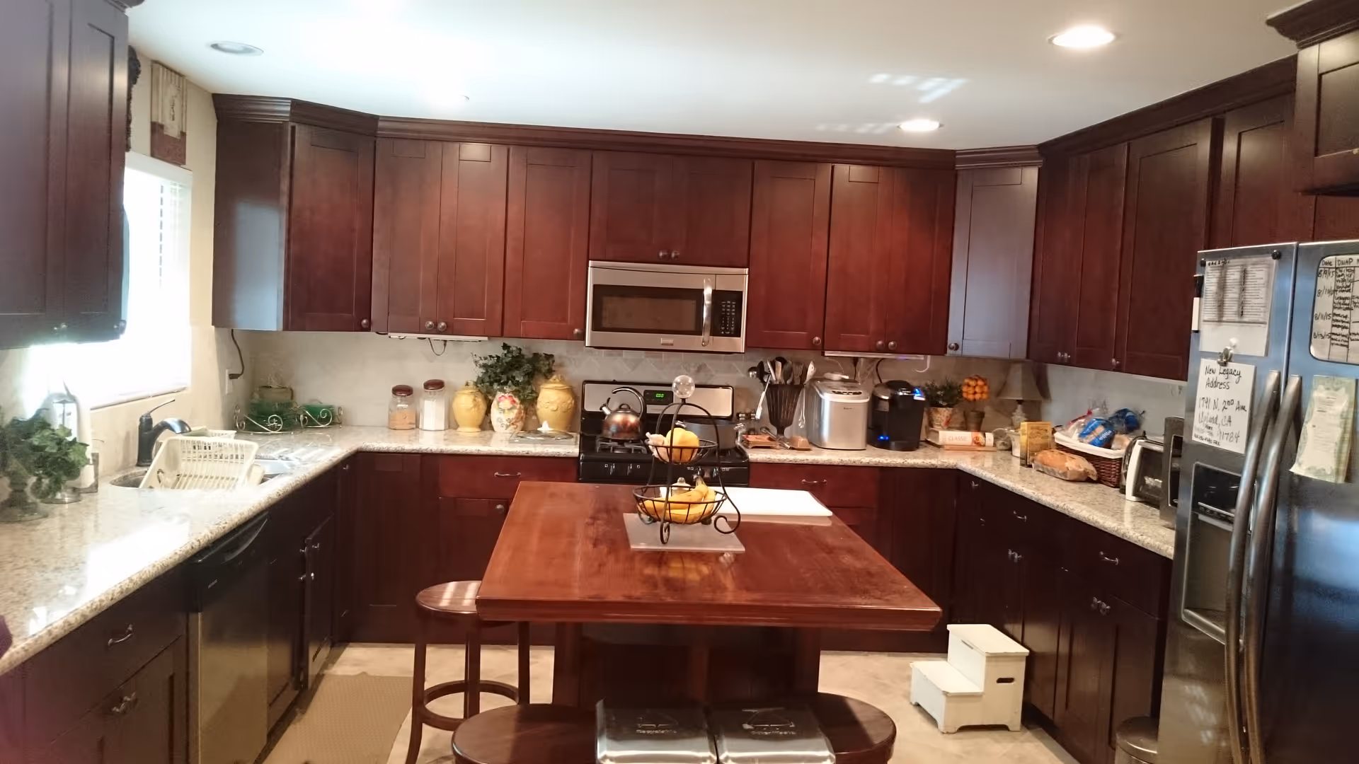 A spacious kitchen with dark wooden cabinets and granite countertops. The kitchen features a central wooden island with a fruit basket on top, a stainless steel microwave above a stove, a dishwasher, a refrigerator with notes attached, and various kitchen appliances and decorative items on the counters.