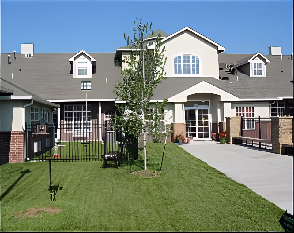 Exterior view of Victoria Falls Skilled Nursing facility showing a large building with multiple windows, a gray roof, and a central entrance with glass doors. There is a green lawn with a few small trees and a paved walkway leading to the entrance under a clear blue sky.