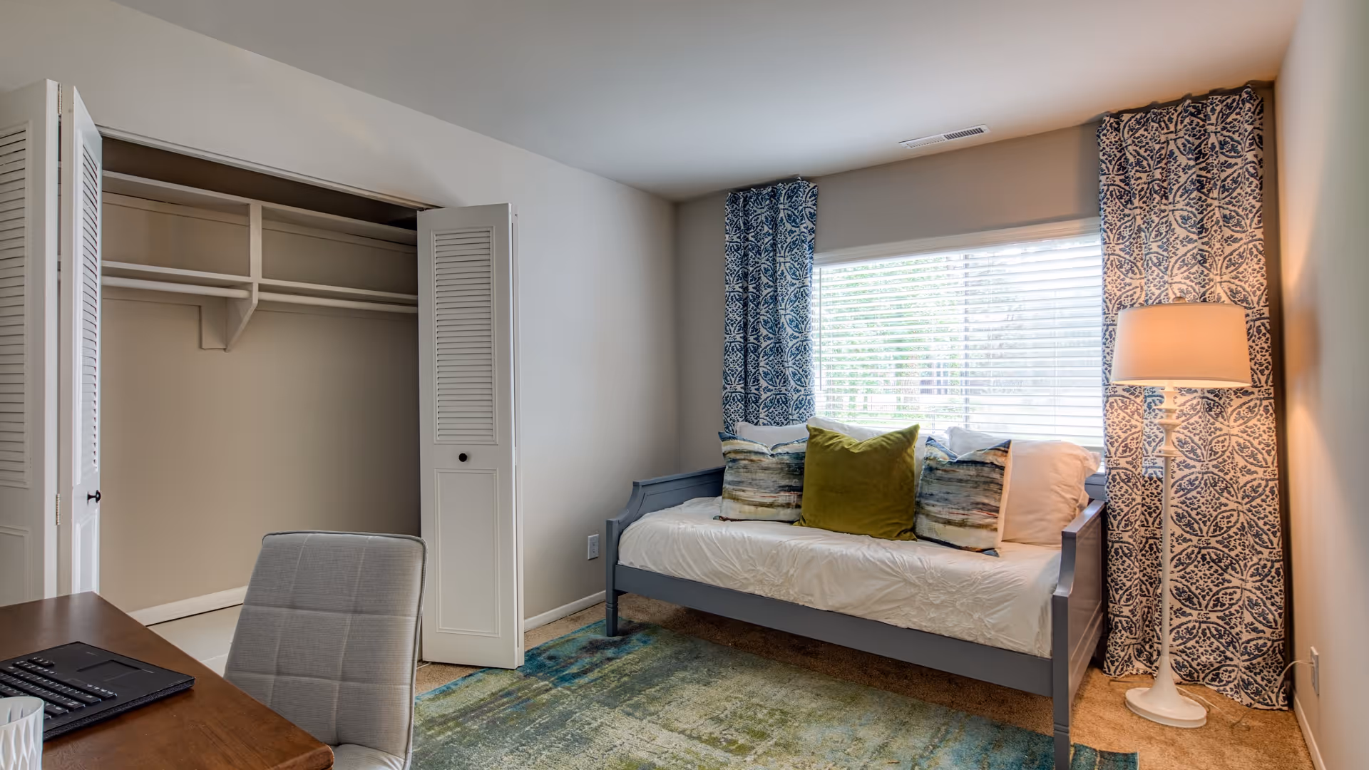 A small bedroom with a daybed featuring white bedding and three decorative pillows in green and multicolor. The room has a large window with blue patterned curtains, a white floor lamp, an open closet with white bi-fold doors, and a wooden desk with a gray upholstered chair.