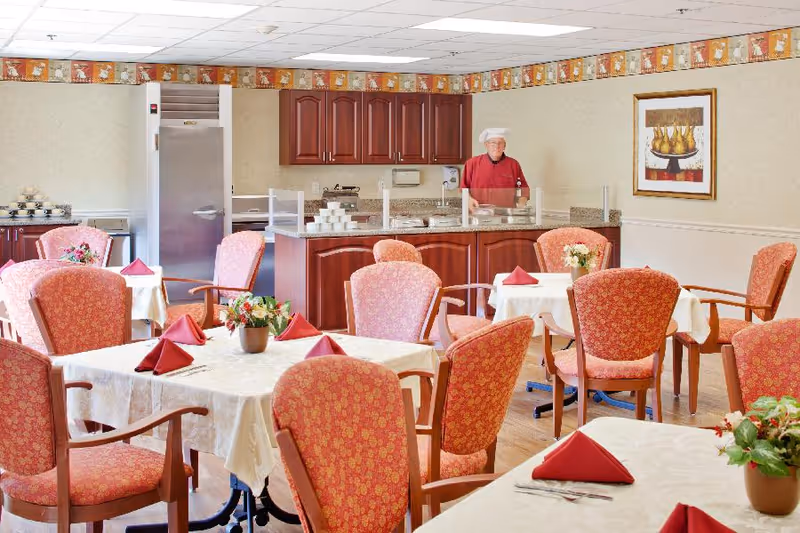 Dining room with several tables covered in white tablecloths and red folded napkins. Each table has a small flower arrangement. There are upholstered chairs with red patterned fabric around the tables. In the background, a staff member wearing a red uniform and white chef hat stands behind a counter with food trays and a protective glass barrier. The room has wooden cabinets, a refrigerator, and a framed painting on the wall.