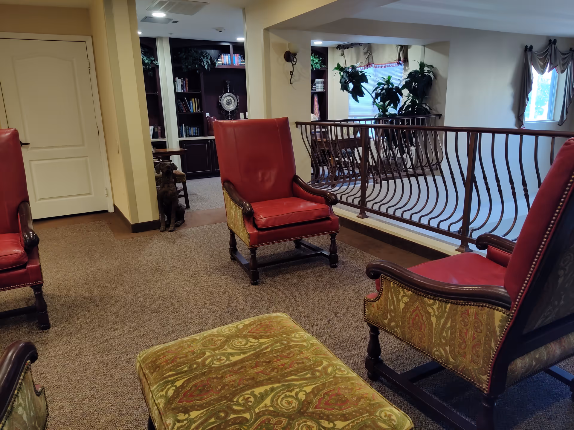 Interior view of a senior living facility lounge area with red leather armchairs and a patterned ottoman on a carpeted floor. A railing overlooks a lower level, and there are plants and a window with curtains in the background. A bookshelf and a decorative dog statue are visible near a doorway.