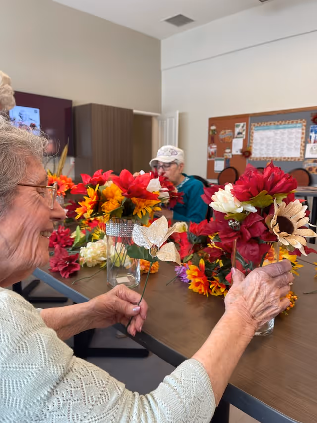 Two elderly women sitting at a table arranging colorful artificial flowers into glass vases in a well-lit room with a bulletin board and chairs in the background.