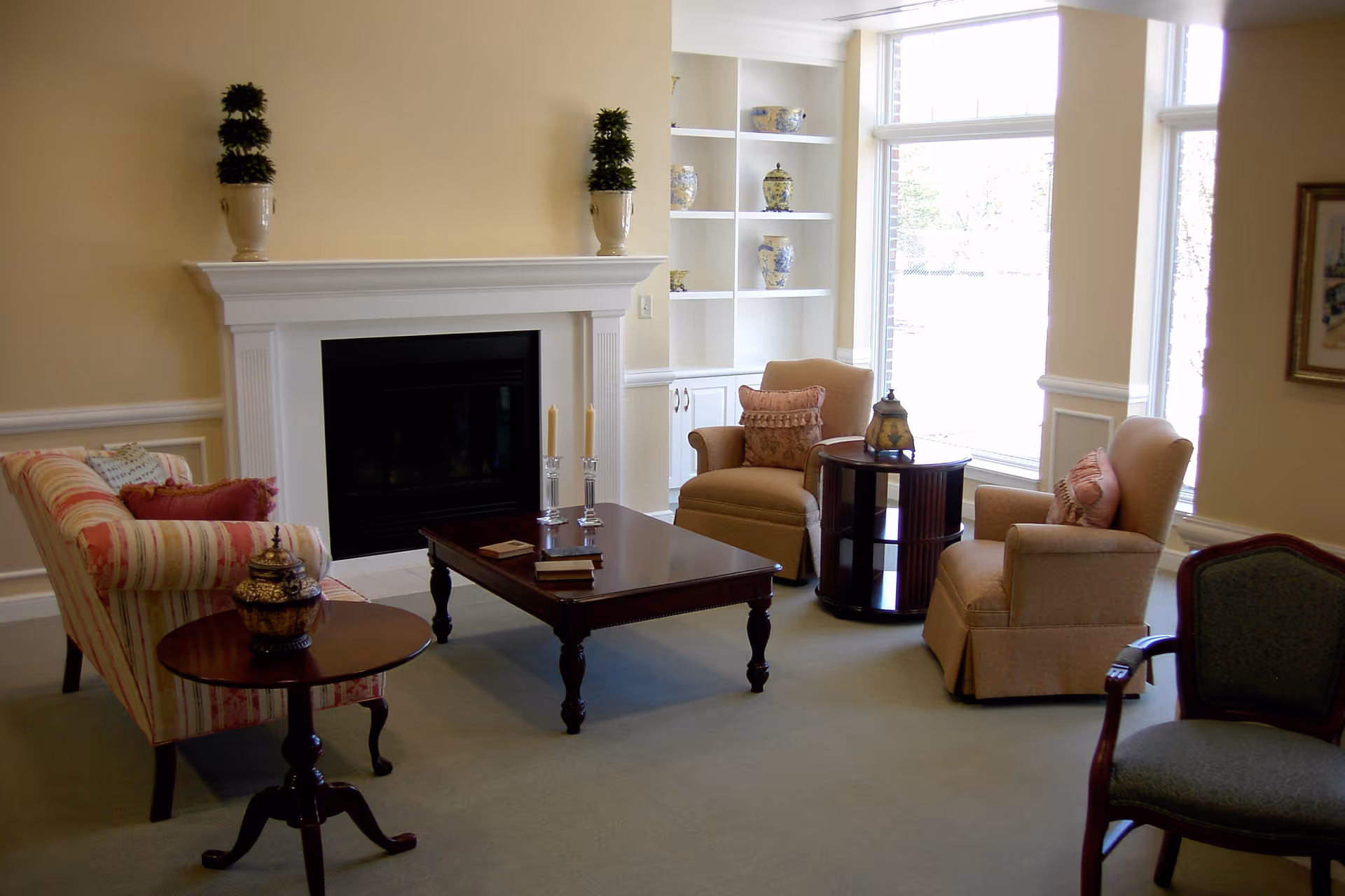 A cozy living room with a fireplace, two beige armchairs with pink cushions, a striped sofa with red and beige pillows, a dark wooden coffee table with candles and books, a round side table with a decorative jar, built-in white shelves with blue and white vases, and large windows letting in natural light.