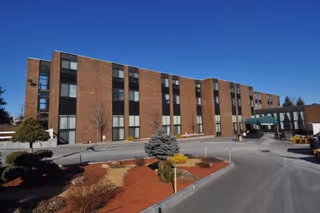 Exterior view of a multi-story brick building with several windows, a driveway, landscaped bushes, and a clear blue sky.