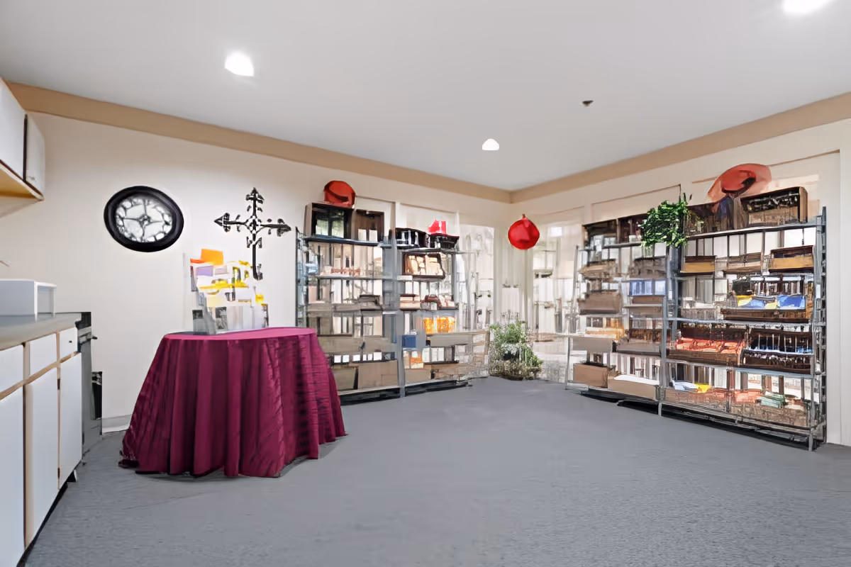 Interior room with carpeted floor, a round table covered with a purple tablecloth, metal shelving units filled with various items, a wall clock, and a decorative metal wall piece. The room has beige walls and ceiling lights.