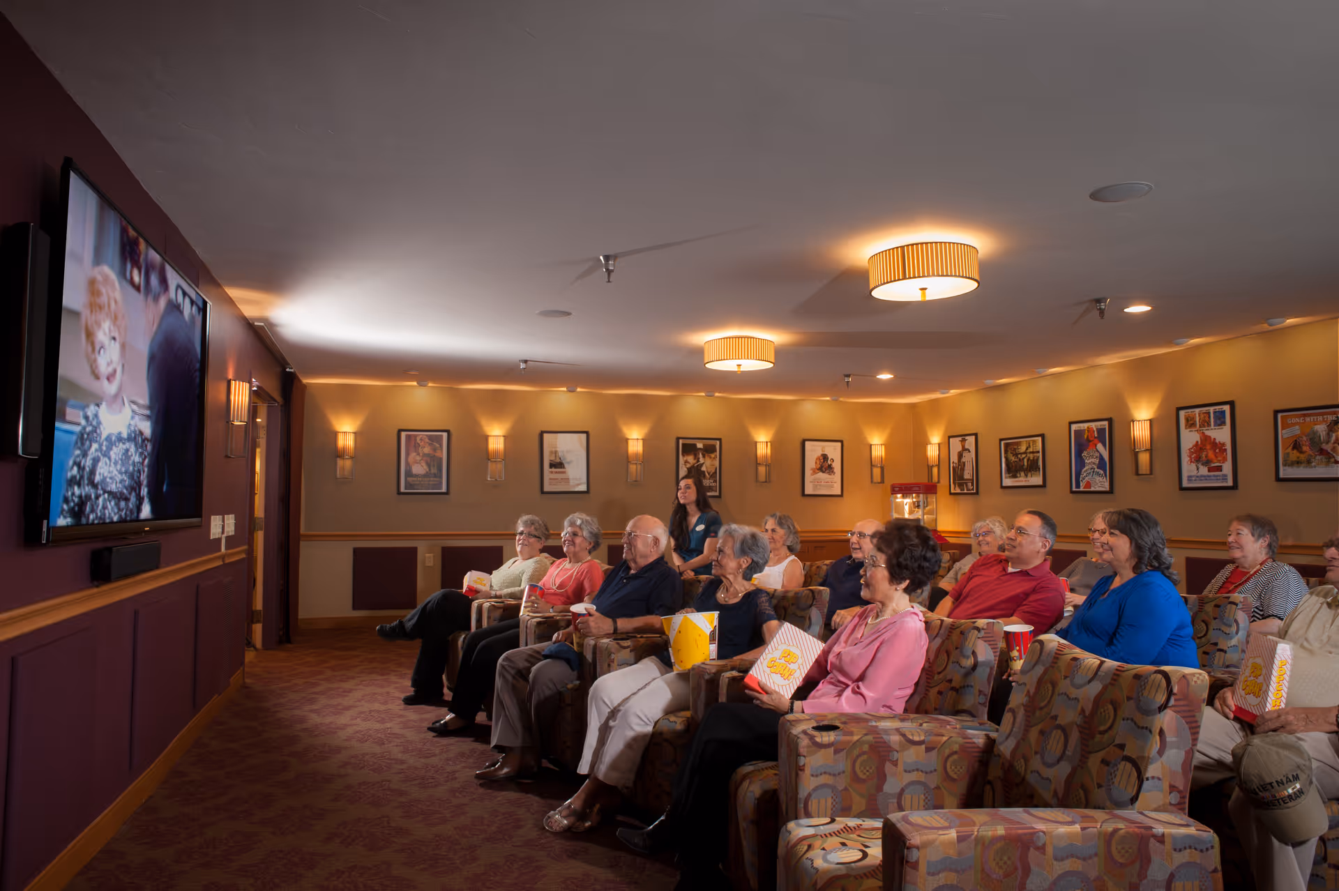 A group of elderly people seated in a cozy movie theater room watching a film on a large screen. They are sitting in patterned armchairs, holding popcorn, with warm lighting and movie posters on the walls.