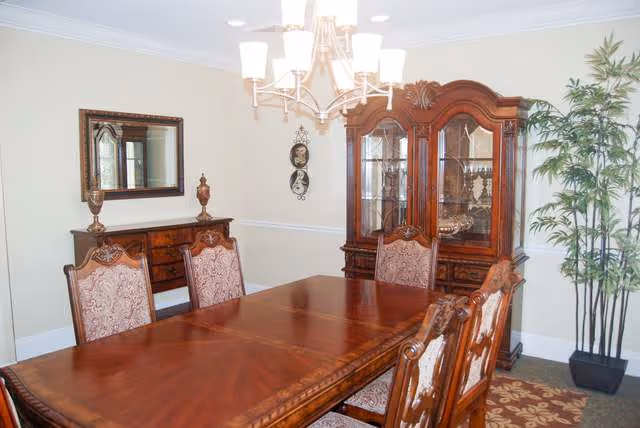 A traditional dining room with a large wooden dining table surrounded by six upholstered chairs. A wooden china cabinet with glass doors is against the wall, displaying decorative items. A sideboard with a mirror above it is also visible. The room is lit by a chandelier and has a potted plant in the corner.