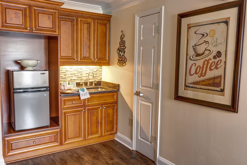 A small kitchenette area with wooden cabinets, a stainless steel mini refrigerator, a granite countertop with a sink, and under-cabinet lighting. A decorative metal wall art of stacked coffee cups is mounted on the wall next to a white door. On the adjacent wall, there is a framed vintage-style coffee poster that reads '5¢ Get more! Coffee Endless cup'. The floor is wooden.