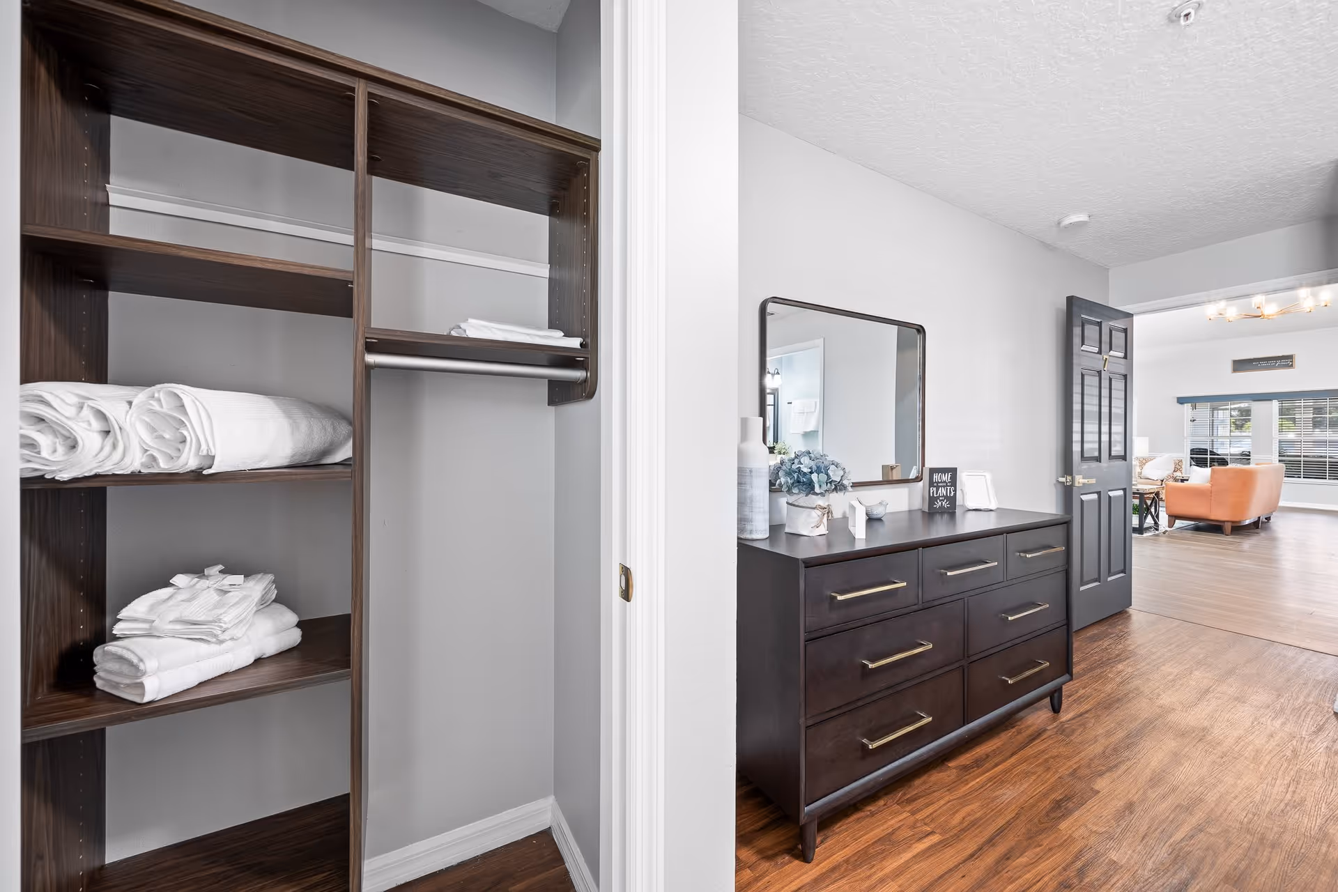 View of a hallway with a dark wooden dresser topped with decorative items and a mirror above it. To the left, there is an open closet with shelves holding neatly folded white towels. The hallway leads to a bright living area with large windows and an orange armchair.