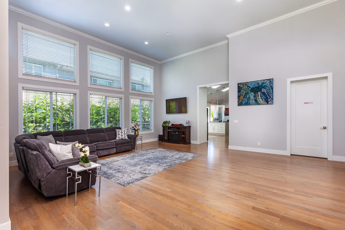 A spacious living room with high ceilings and large windows covered with blinds, allowing natural light to fill the room. There is a large gray sectional sofa with pillows, a small side table with a flower arrangement, a blue patterned area rug on the wooden floor, a wall-mounted flat-screen TV above a wooden cabinet, and a colorful abstract painting on the wall near a white door. The room opens into a kitchen area visible through a doorway.