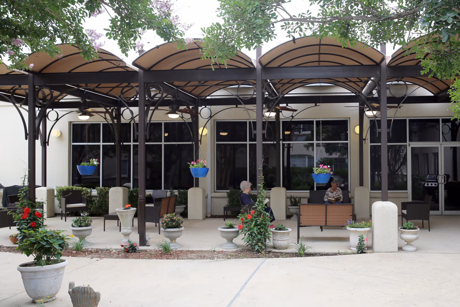 Outdoor covered patio area with seating and potted plants. Two elderly people are sitting and conversing under the canopy structure with hanging flower pots. The background shows large windows and doors of the building.