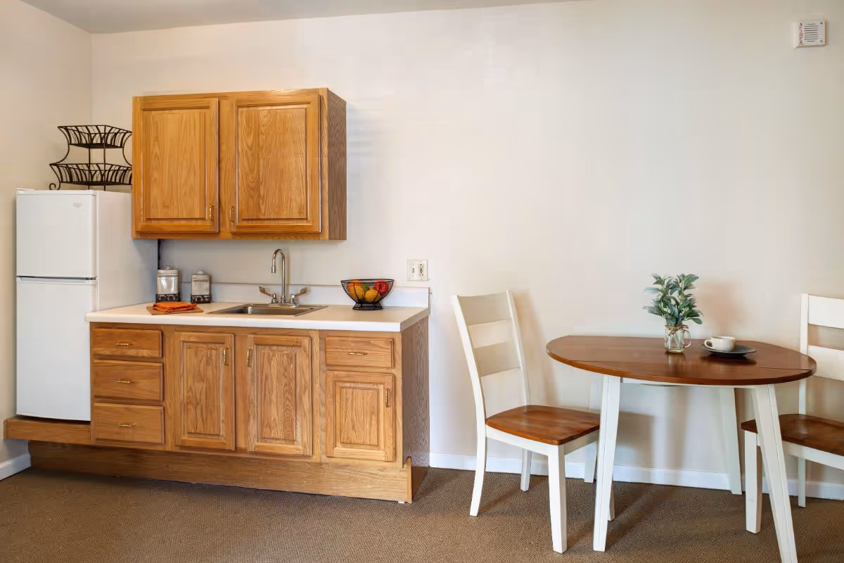 Small kitchenette with wooden cabinets, a sink and refrigerator next to a round dining table with two chairs.