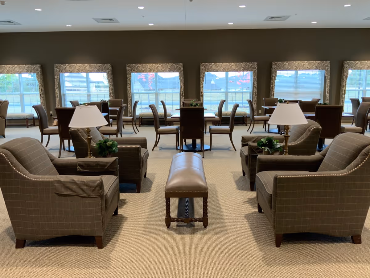 A spacious and well-lit common area with multiple seating arrangements including armchairs, small tables with chairs, and a long bench in the center. The room has large windows with patterned curtains allowing natural light to enter, and two table lamps on side tables beside the armchairs. The carpeted floor and neutral color palette create a warm and inviting atmosphere.
