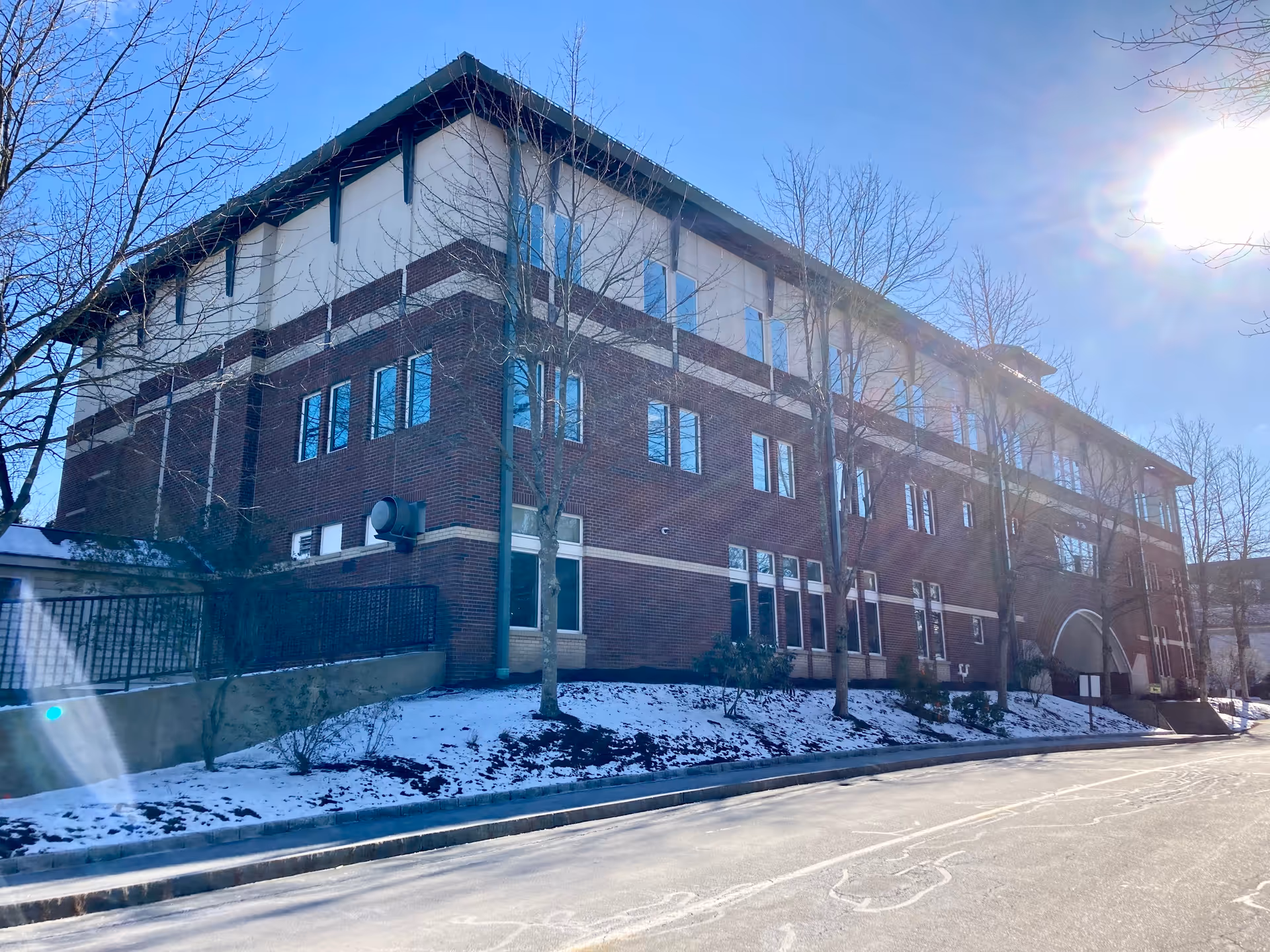 Exterior view of a large brick building with multiple windows, leafless trees, and a light dusting of snow on the ground under a clear blue sky with the sun shining brightly.