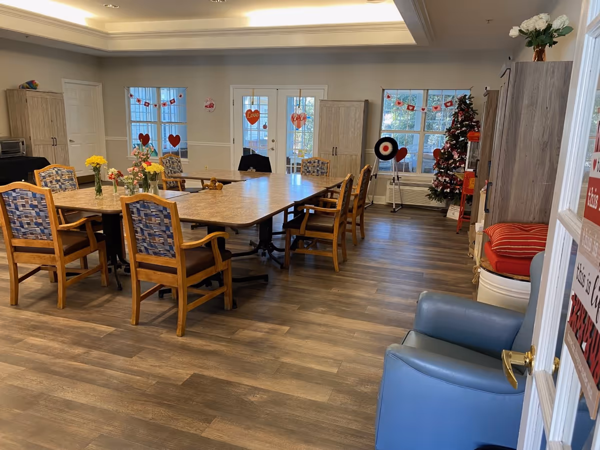 A well-lit communal dining room with wooden tables and cushioned chairs arranged around them. The room has wood flooring, two windows, and a glass door decorated with heart-shaped Valentine’s Day decorations. There are cabinets along the walls, a Christmas tree in the corner, and several vases with flowers on the tables. A blue armchair is visible near the entrance.