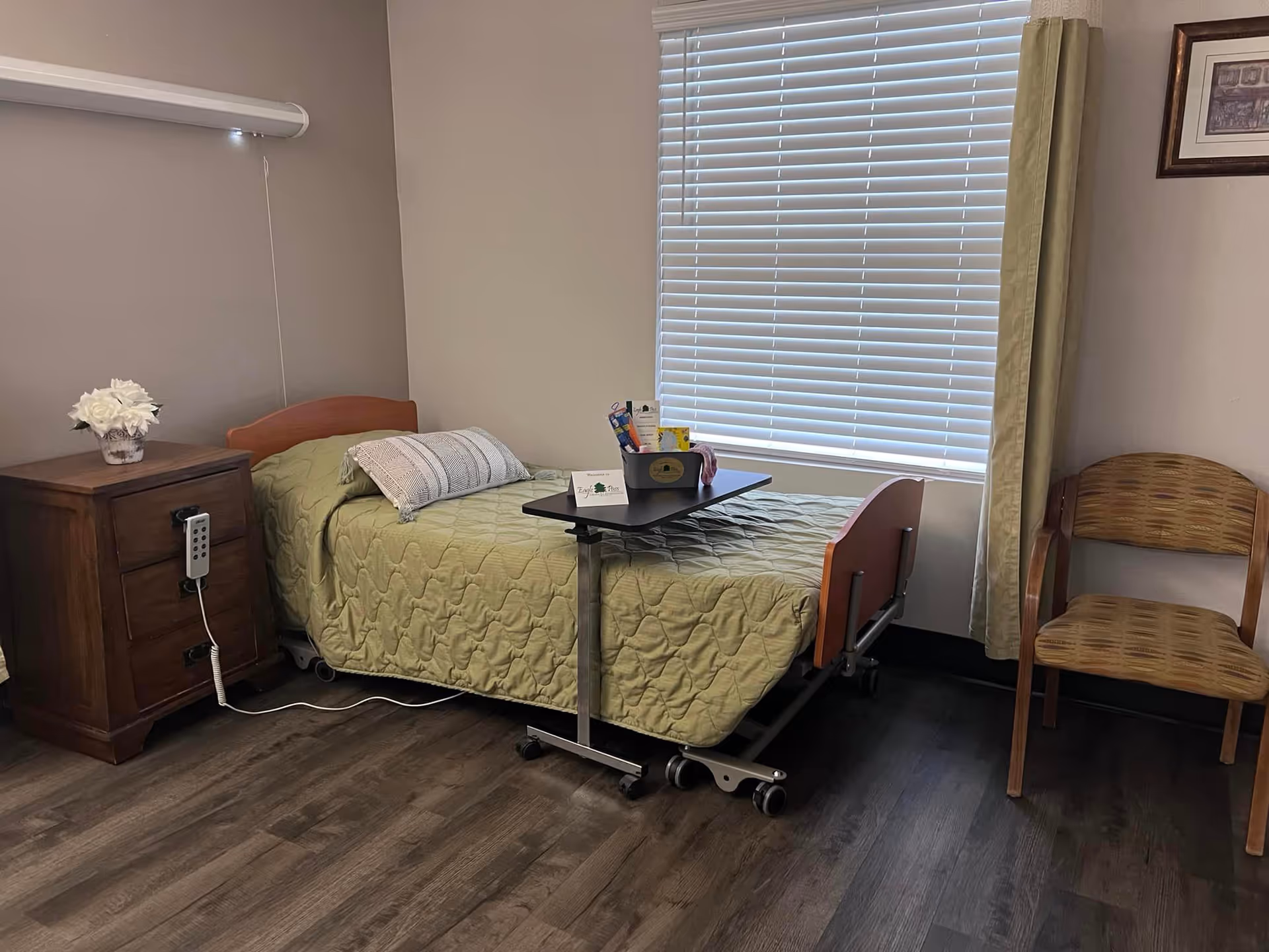 A small nursing facility bedroom with a single hospital-style bed, overbed table, wooden nightstand with flowers, and a chair beside a window with blinds.