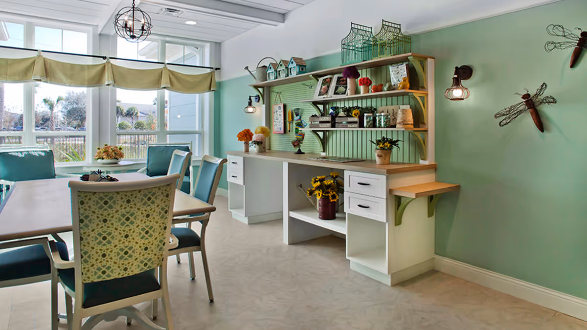 A bright and airy room with large windows letting in natural light. The room features a table with several chairs around it, some with patterned backs and teal cushions. Against one wall is a white and wood craft station with shelves holding decorative items, small birdhouses, jars, and flowers. The walls are painted a soft green, and there are two decorative dragonfly wall hangings. The floor is a light-colored tile, and a modern pendant light hangs from the ceiling.