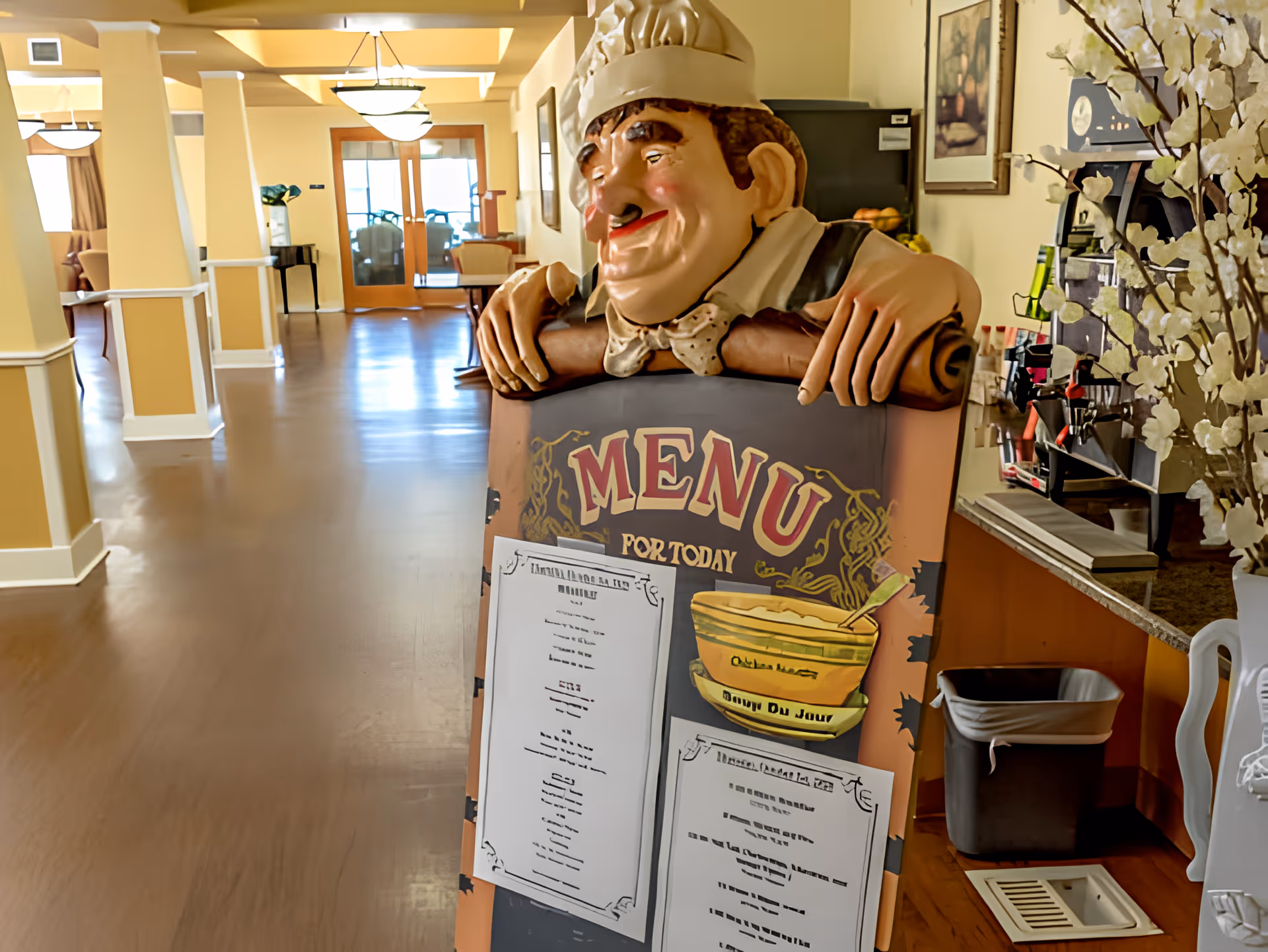 A hallway in a senior living facility with a decorative chef statue holding a menu board displaying the daily menu. The hallway has beige walls, wooden flooring, and ceiling lights. There is a trash bin and a coffee station on the right side, and seating areas visible in the background.