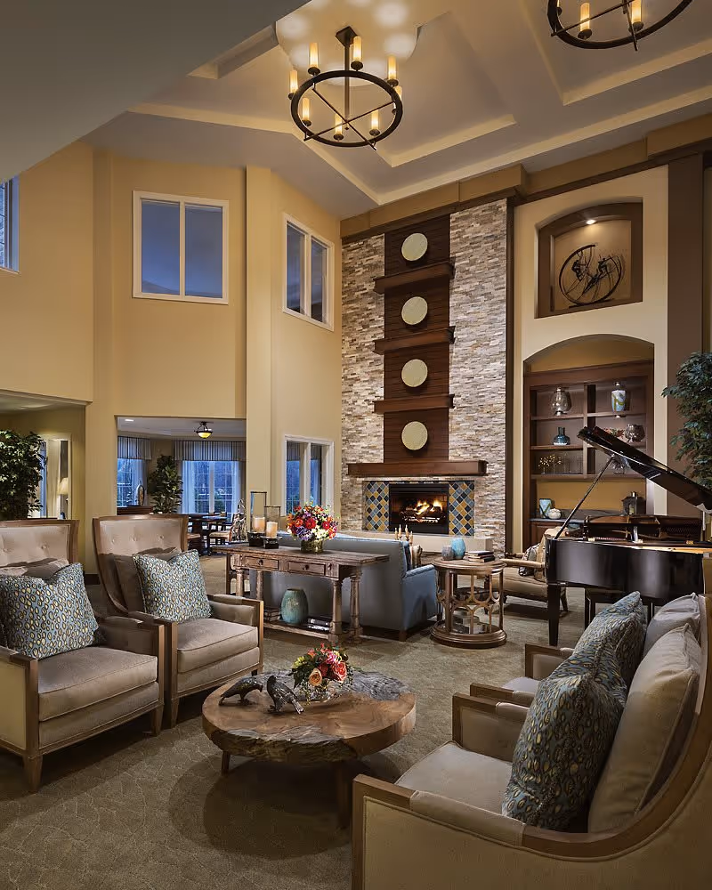A spacious and elegant living room area with high ceilings, featuring a stone fireplace with decorative circular accents, comfortable armchairs with patterned cushions, a wooden coffee table with floral arrangements, a grand piano, and built-in shelves with decorative items. Large windows and warm lighting create a cozy atmosphere.
