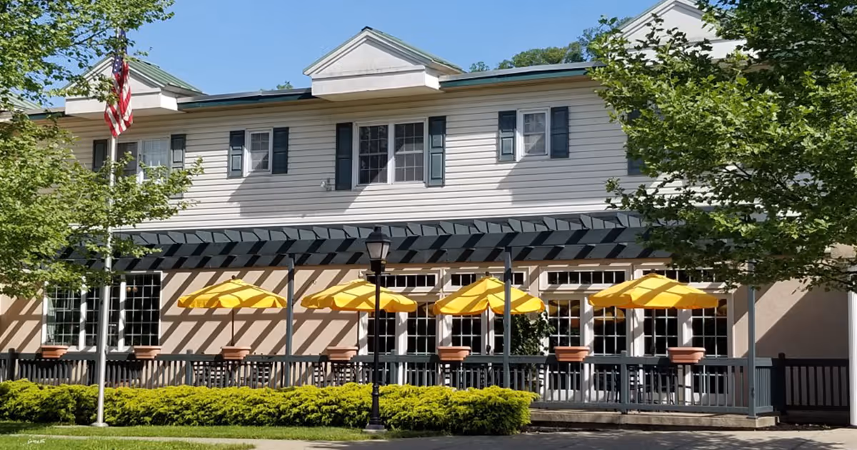 Exterior view of a senior living facility building with a covered patio area featuring several yellow umbrellas over outdoor tables and chairs. The building has white siding, green shutters, and a flagpole with an American flag. Trees and shrubs surround the patio area.