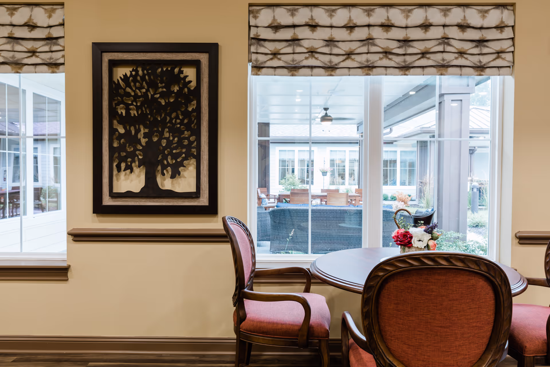 A cozy interior seating area with a round wooden table and four upholstered chairs with reddish fabric. On the table is a small floral arrangement in a basket. Behind the table are large windows with patterned Roman shades, offering a view of an outdoor patio area with wicker furniture and a ceiling fan. A framed metal tree wall art hangs on the beige wall between the windows.