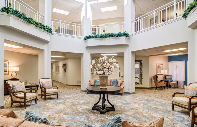 Atrium-style interior common lounge with seating surrounding a central round table topped with a floral arrangement and upper-level balcony railings.