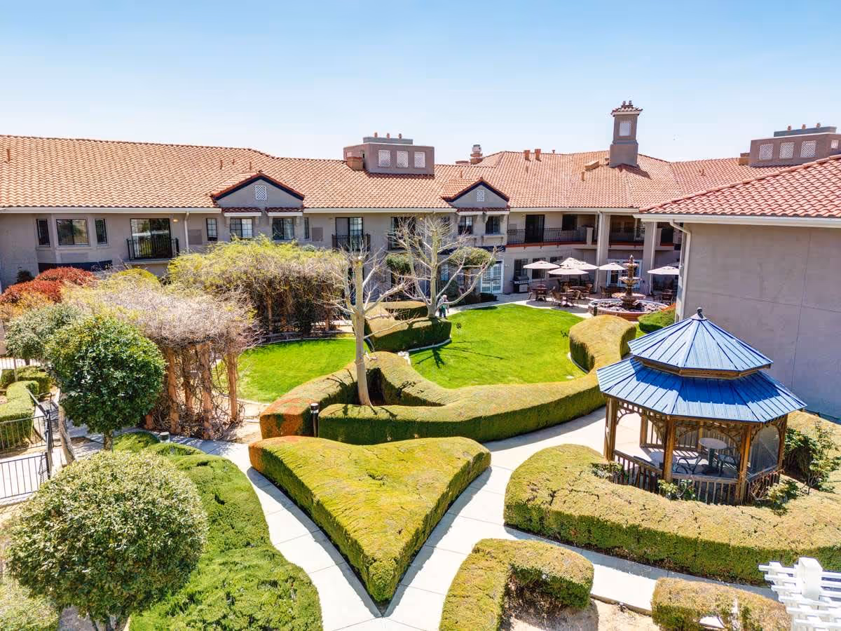 Outdoor courtyard of Whispering Winds of Apple Valley featuring neatly trimmed hedges, a green lawn, a wooden gazebo with a blue roof, and a building with a red tile roof surrounding the area. There are tables with umbrellas near the building and a clear blue sky overhead.