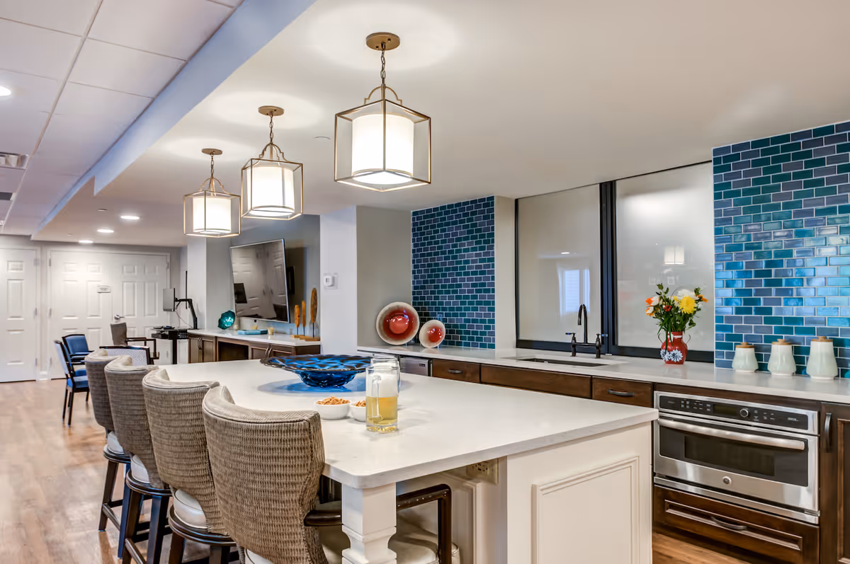 Modern kitchen area with a large white island countertop surrounded by four wicker bar stools. Above the island hang three pendant lights with square shades. The back wall features a blue and green tiled backsplash, a sink with a black faucet, a built-in oven, and decorative items including a vase with flowers and ceramic containers. In the background, there is a TV mounted on the wall and additional seating.