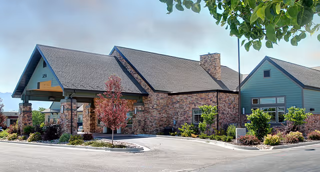 Exterior view of a single-story building with stone and blue siding, a covered entrance supported by stone pillars, surrounded by landscaped greenery and trees under a clear sky.