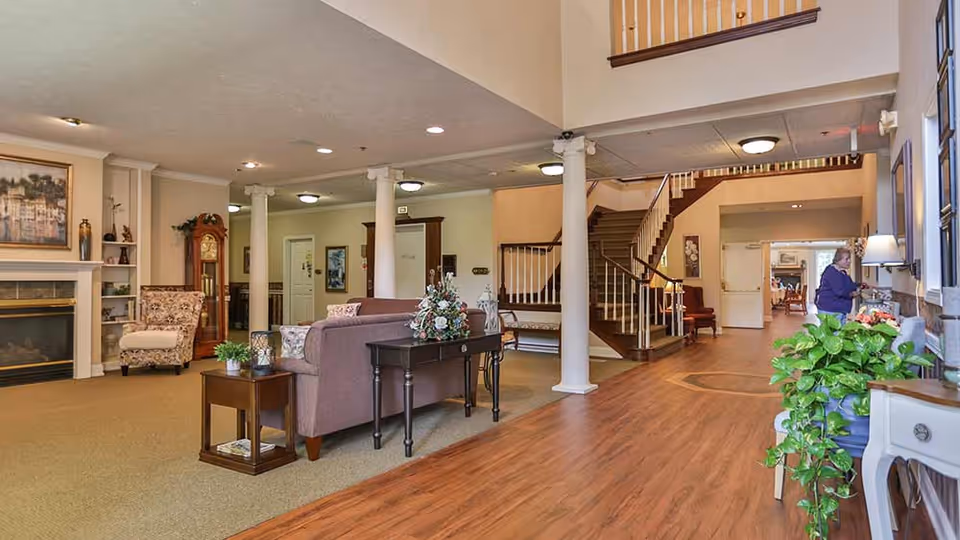 Spacious senior living facility common area with a seating arrangement including a sofa and armchair near a fireplace. The room features wooden flooring, white columns, a staircase leading to an upper floor, and various decorative plants and artwork. A person is seen near the window on the right side of the image.