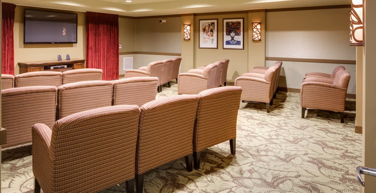 Small screening/media room with rows of upholstered chairs facing a wall-mounted TV and movie posters.