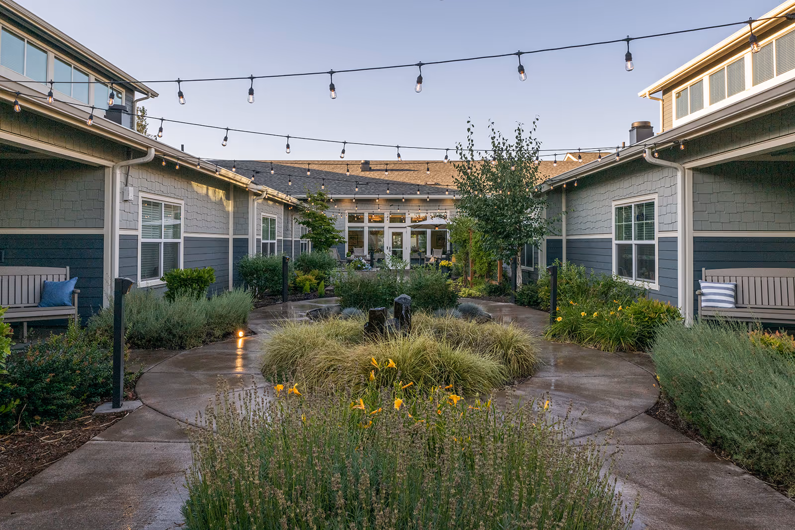 Outdoor courtyard area at Battle Creek Memory Care with a circular pathway surrounded by greenery and flowers. Benches with cushions are placed along the sides near the building walls. String lights are hung overhead, creating a cozy atmosphere.