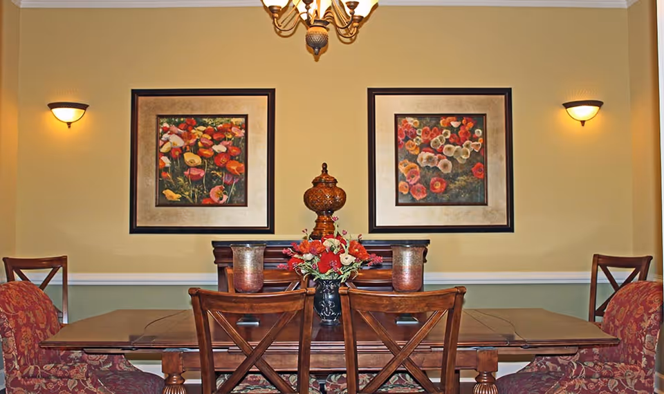 A dining room with a wooden table surrounded by six chairs, two of which have red patterned upholstery. On the table is a floral centerpiece and two decorative candle holders. The wall behind the table is painted yellow and green with white trim and features two framed floral paintings. Two wall sconces provide lighting, and a chandelier hangs from the ceiling above the table.