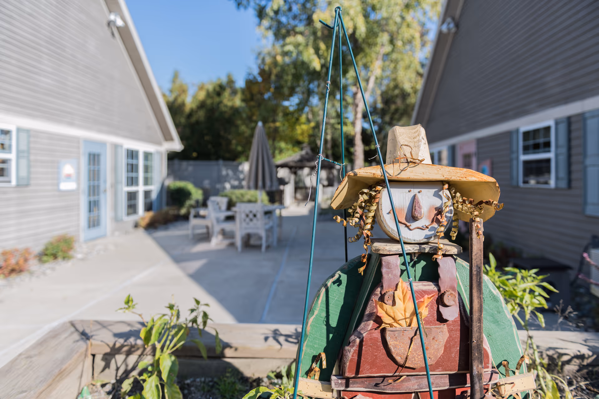A close-up of a decorative scarecrow figure with a hat and curly hair made of metal and wood, placed in a garden bed. In the background, there is an outdoor patio area with tables, chairs, and umbrellas between two buildings with gray siding and blue shutters under a clear blue sky.