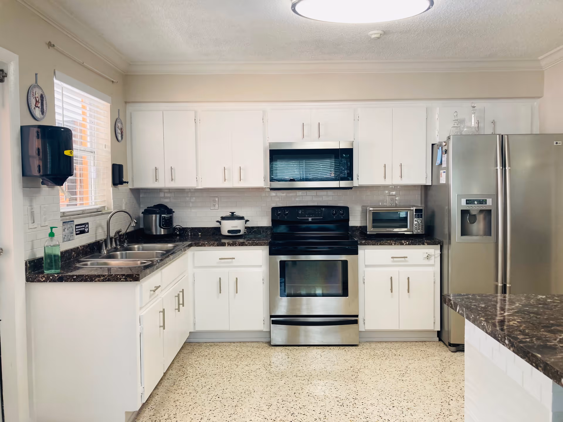 A clean and modern kitchen with white cabinets, a stainless steel stove and microwave, a stainless steel refrigerator with water dispenser, a double sink under a window with blinds, a toaster oven, a rice cooker, and a slow cooker on the dark granite countertops. The floor is light-colored with a speckled pattern.