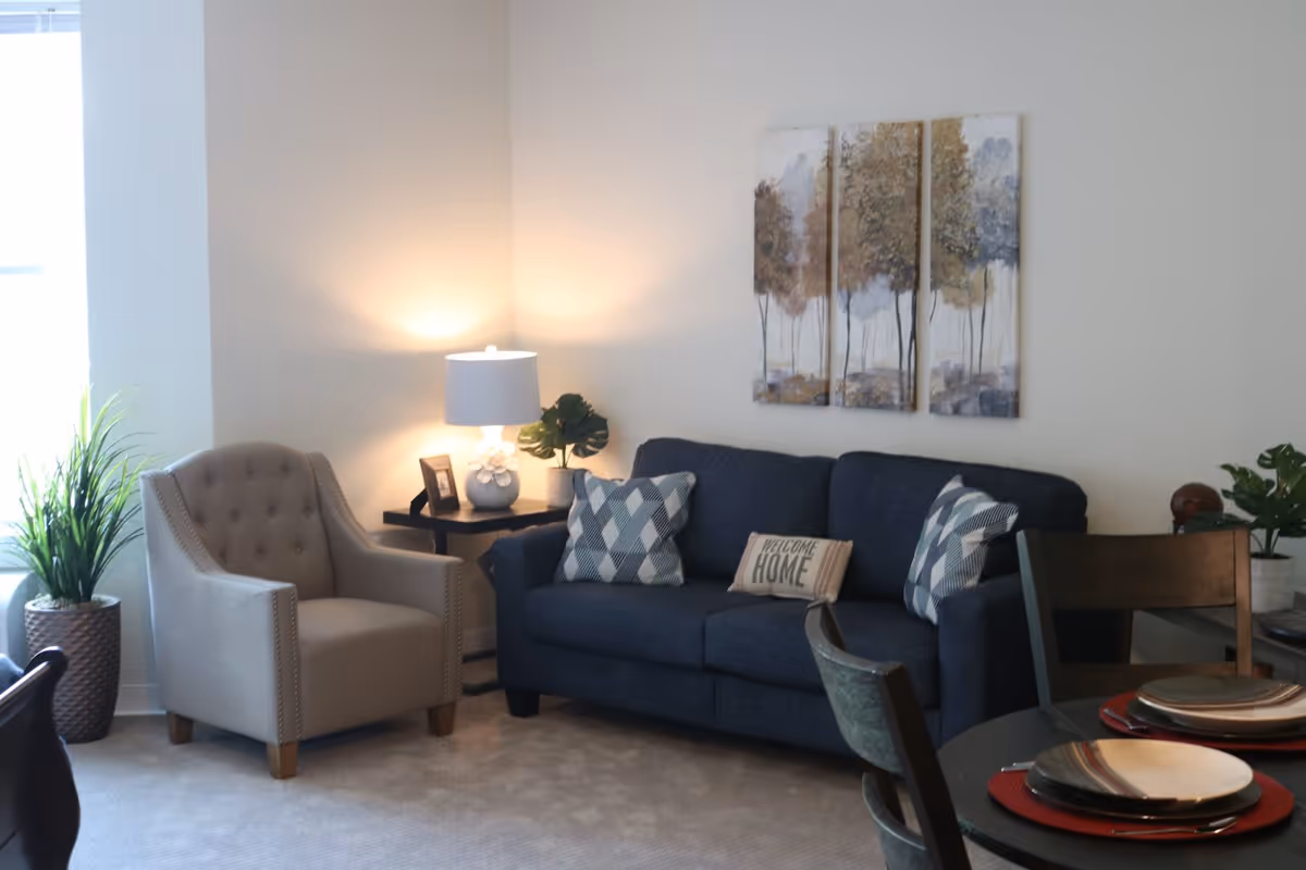 A cozy living room area with a blue sofa adorned with patterned pillows and a 'Welcome Home' pillow, a beige armchair, a side table with a lamp and a small plant, a three-panel wall art depicting trees, and a dining table set with plates and placemats.