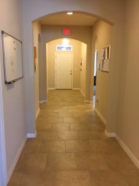 A clean, well-lit hallway with beige tiled flooring and light-colored walls. There are two archways on the left and right sides of the hallway, a whiteboard mounted on the left wall, and bulletin boards on the right wall. At the end of the hallway is a white door with a window above it and an illuminated red exit sign above the door.