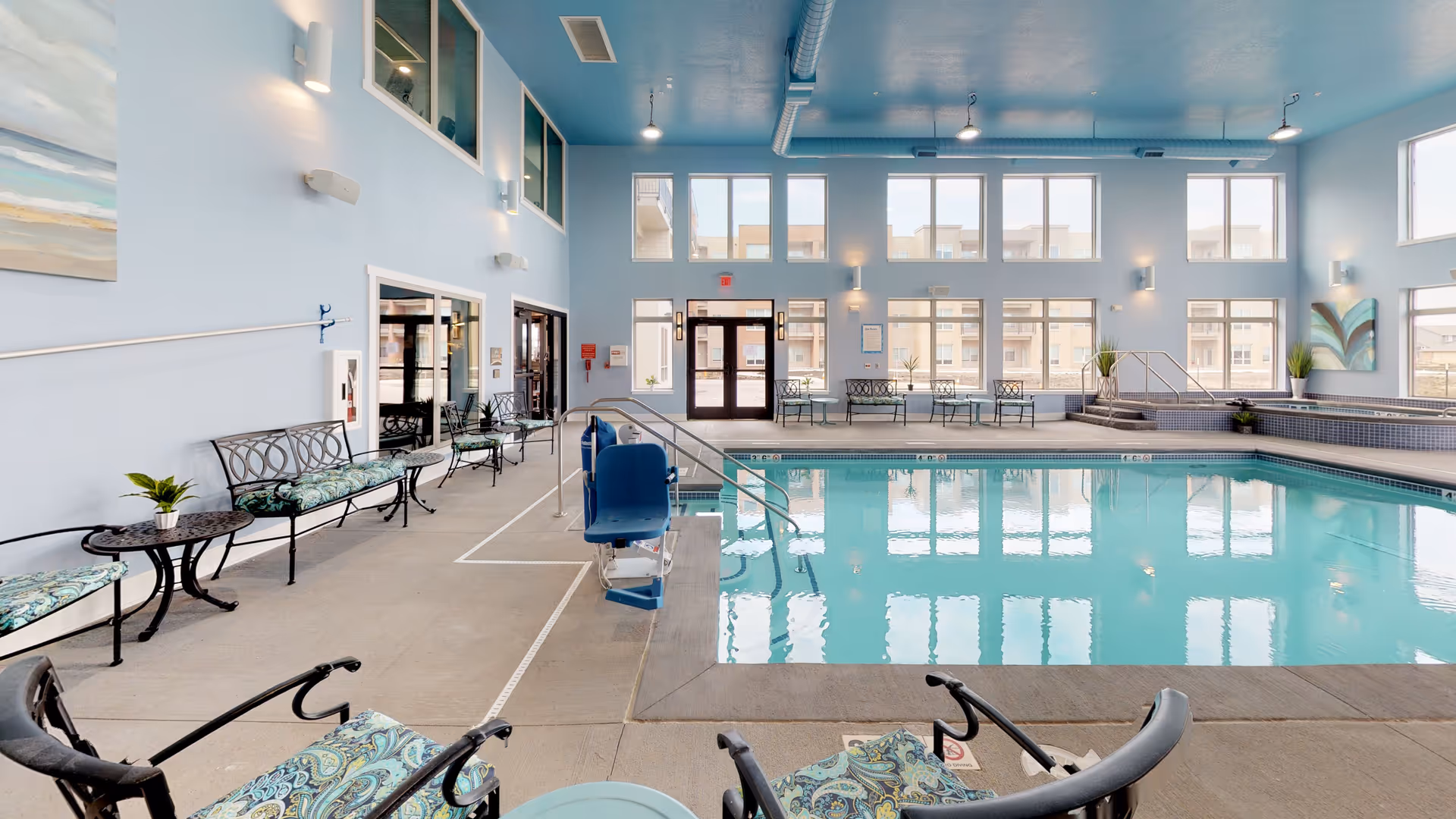 Indoor swimming pool area with light blue walls and ceiling, large windows letting in natural light, several cushioned benches and chairs with patterned upholstery around the pool, a pool lift for accessibility, and potted plants placed near the walls.